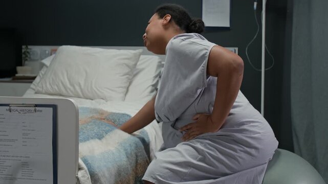 Pregnant Black woman sitting on fitness ball in hospital room,holding belly during labor contractions. Footage is ideal for maternity care and childbirth education, side view shot