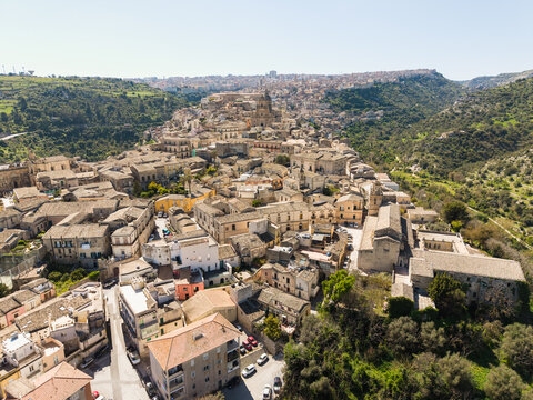 Aerial view of the historic baroque architecture and stone buildings of Ragusa Ibla nestled between green valleys under a clear sky in Ragusa, Sicily, Italy.