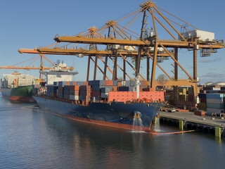 Naklejka premium container ship with water from anchor moored at container terminal. cargo operation in progress. big overhead type cranes working