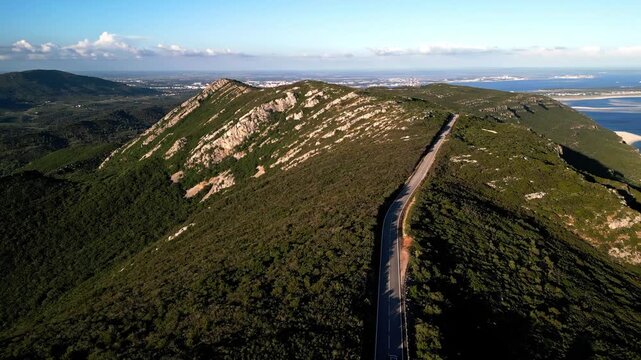 Drone footage of a winding highway along the Arr&aacute;bida Natural Park ridge in Set&uacute;bal, Portugal, showcasing lush green hills and a distant ocean coastline