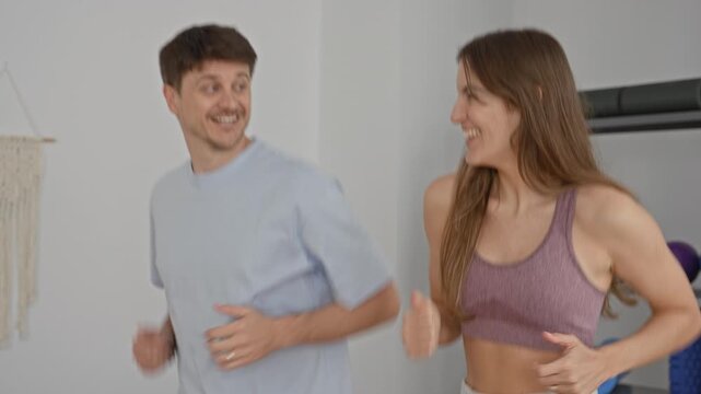 Young man and woman jogging side by side in a fitness studio, woman bare midriff, hands pumping, yoga mats and foam roller on shelves visible; shared joy and connection.