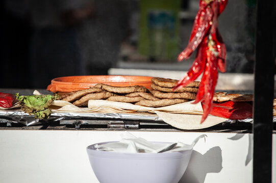 Pljeskavica patties cooking on a rostilj stand in a market, showing Balkan grilled meat prepared on flatbread with peppers, foil and equipment for takeaway customers at the counter.