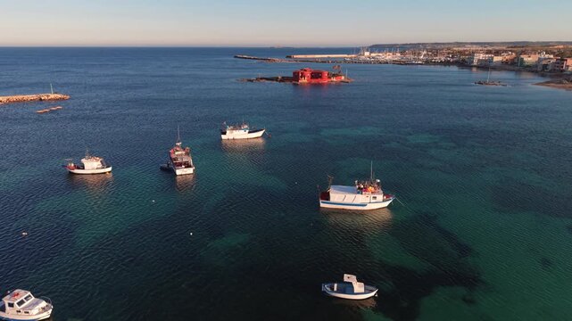 Aerial view of fishing boats floating on clear turquoise water near the red villa of Isolotto Brancati during golden hour in Marzamemi, Sicilia, Italy.