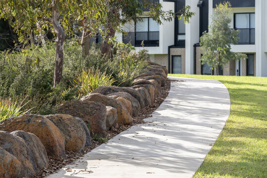 A curved concrete footpath bordered by a row of large natural boulders used as landscape edging, separating the pathway from a native garden bed