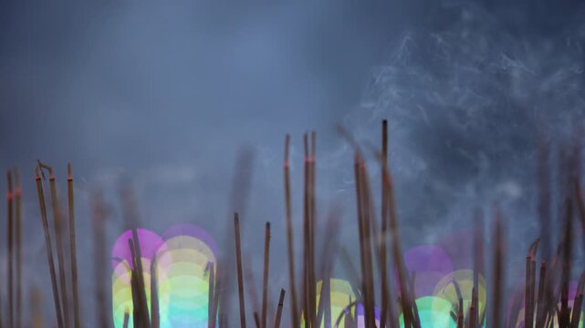Cinematic incense smoke rising softly from altar against colorful festival bokeh lights background. Continuous flow of white smoky texture with close up focus on burning joss sticks.