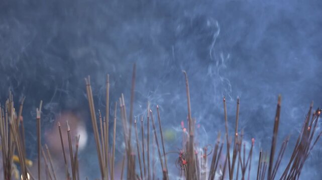 Close up of burning incense sticks bundle with thick white smoke rising full frame. Active smoky texture with blurred silhouette of worshippers walking by during night ritual b-roll.
