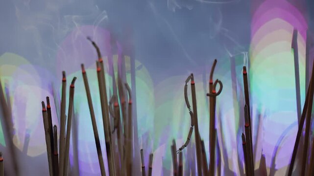 Close up of burning incense sticks bundle with thick white smoke rising full frame. Active smoky texture against vibrant colorful festival bokeh lights background b-roll.
