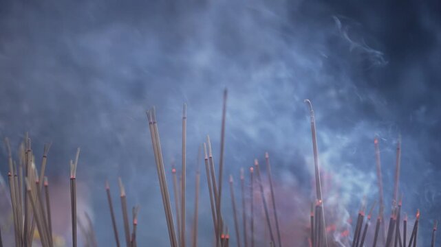 Cinematic incense smoke rising full frame from burning joss sticks bundle. Active smoky texture with blurred silhouette of worshippers praying against dark night sky background.