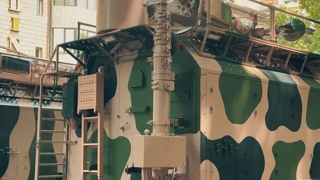 This heavy armored military camouflage truck remains parked on a sunny city street during an outdoor public exhibition featuring modern tactical defense vehicles.