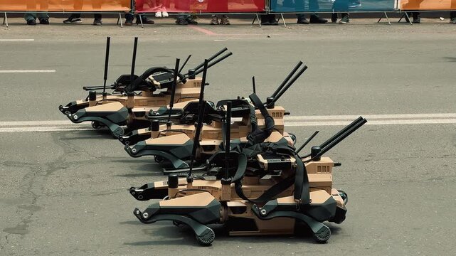 Four quadruped military robotic dogs sit lined up on a city street while police officers and soldiers walk past during an outdoor public technology and defense exhibition event.

