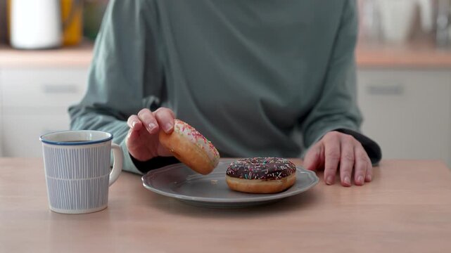Woman having unhealthy lunch at home, sitting in kitchen takes doughnuts from plate but refusing to eat, female practicing healthy eating and willpower, reducing sugar intake, taking care of health