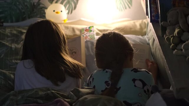 Bookworm twins reading bedtime stories together in bed by night lamp, rear view