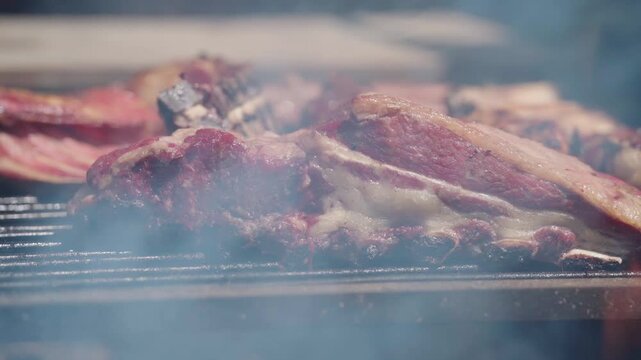 Close-up view of traditional Argentine asado de tira being turned with metal tongs over a smoky wood fire grill, showcasing authentic rustic barbecue cooking and juicy grilled beef ribs.