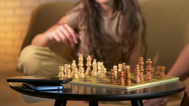 Young girl playing chess with an adult. Smart young girl with long hair concentrating on a chess game, making strategic moves on the chessboard while playing with an adult putting aside smartphones