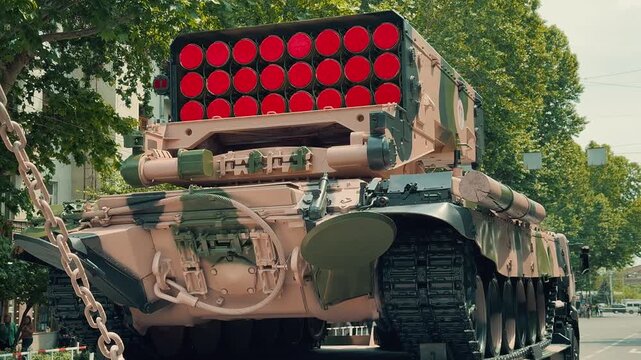 Two camouflage TOS-1A multiple rocket launcher tanks are displayed on a heavy transport flatbed trailer on a city street during a public national army defense parade exhibition event.