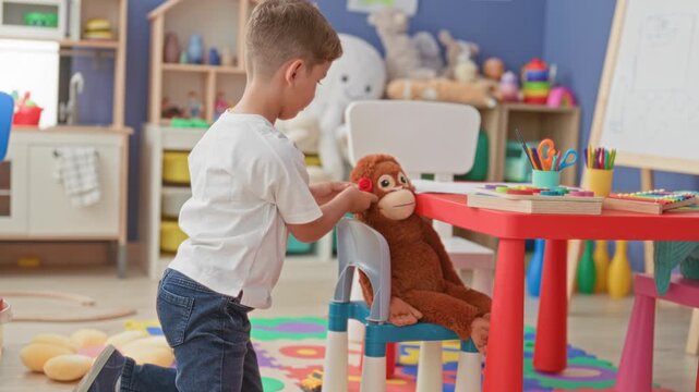Boy kneels and manipulates a red toy beside a seated brown stuffed monkey at a small red table amid colorful toys and shelves in a building; playful curiosity.