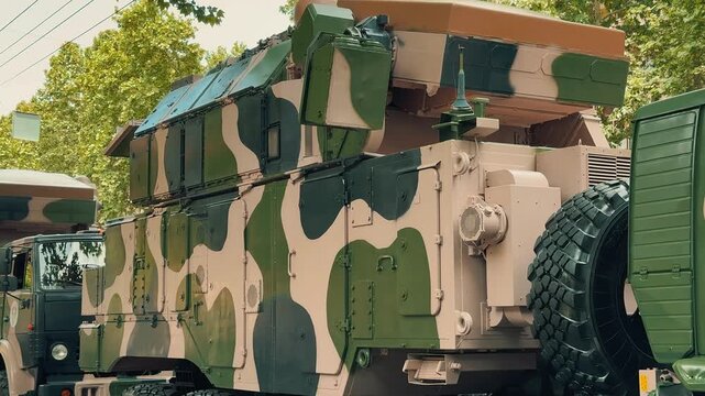 A large camouflage military support and communication truck is parked on a city street during a public national army defense parade and exhibition event showcasing specialized heavy equipment.

