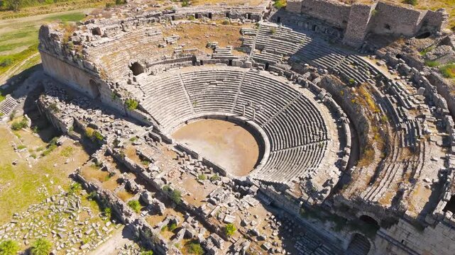 Balat, Turkey. Ancient Theatre of Miletus, sweeping semicircular stone cavea with radial stairways and arched entrances, aerial view over extensive archaeological ruins under bright summer sun. Aeria