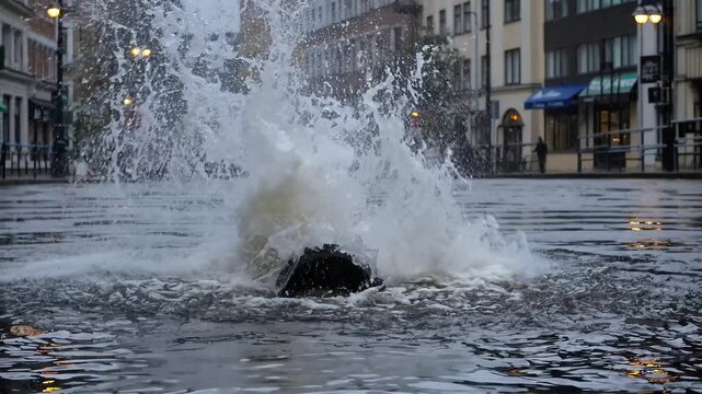 Exploding dirty water pipe bursts out from open drain hole on flooded city square after storm, rapid sewer geyser breaks street pavement tiles and creates emergency situation on avenue road