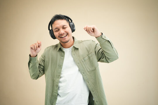 Excited young Asian man wearing headphones dancing with arms raised, laughing and enjoying music in casual olive green shirt and studio setting