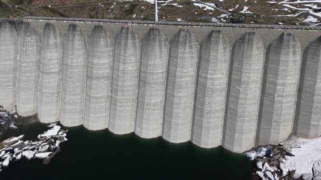 Aerial view of the massive concrete Lucendro Dam with dark water and patches of melting snow on the rocky mountain slopes in Airolo, Ticino, Switzerland.