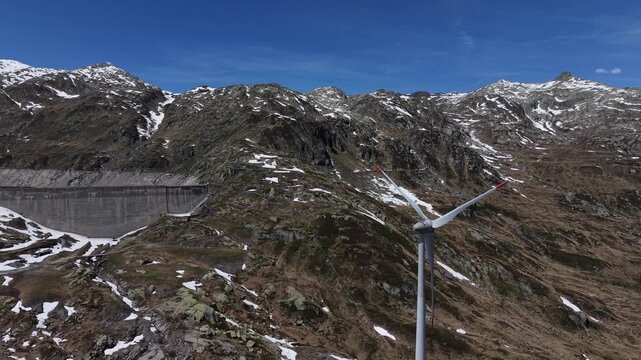 Aerial view of the Centrale idroelettrica di Lucendro dam and a wind turbine among snow-capped mountains under a clear blue sky in Airolo, Ticino, Switzerland.