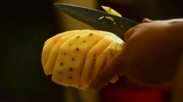 Hands slice a pineapple with care. Golden flesh gleams under warm light. Knife glides through fruit with precision. Fruit's pattern reveals juicy interior. Scene suggests kitchen preparation
