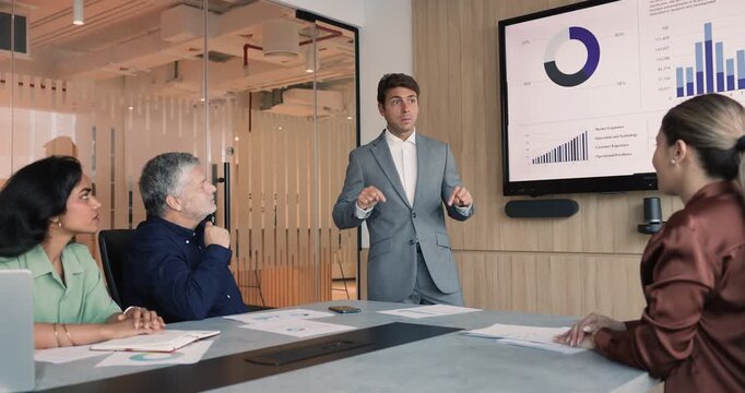 Confident young Latin businessman presenting project KPI benchmarks, standing at meeting table in boardroom, talking to diverse team of coworkers, offering plan and strategy of development