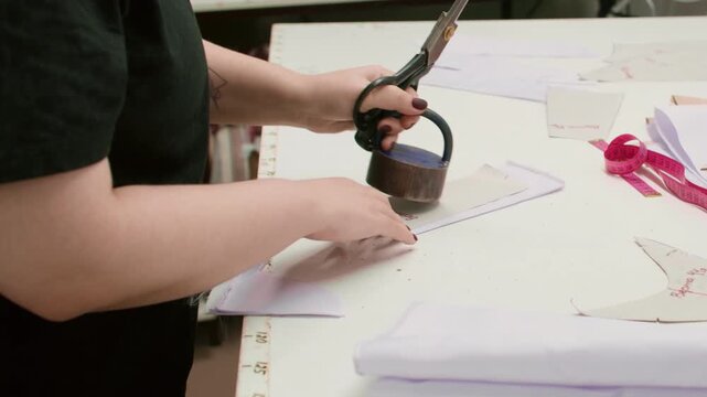 Seamstress occupation. Tailor desk. Woman hand cutting curved garment textile pattern with heavy weight scissors in studio interior.