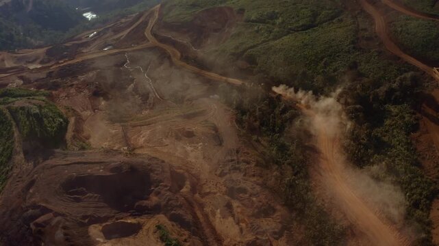 Top-down aerial tracking shot of an active iron ore mine, Red laterite soil and mining equipment contrast with surrounding green forest and mountains in Xieng Khouang province, Lao PDR.