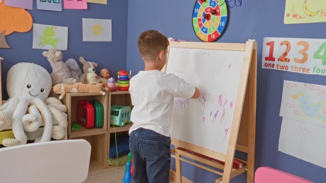 Boy standing erases easel whiteboard with red eraser in preschool classroom surrounded by stuffed toys and posters; concentration learning.