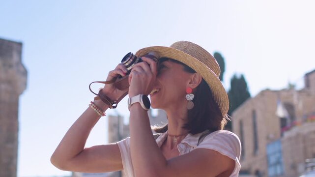 A young woman in a straw hat and summer dress holds the camera in front of her face, looking up at a sunlit historic town square. She stands in the center of the frame