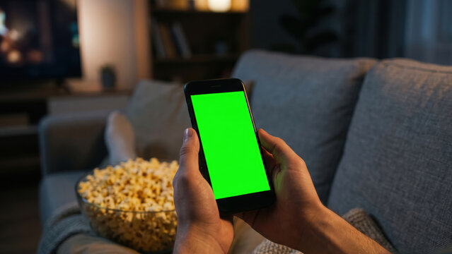 Relaxed POV holding a green screen mobile phone on a couch with a bowl of out-of-focus popcorn. Excellent for food delivery apps, movie streaming UI mockups, and cozy home entertainment layouts.