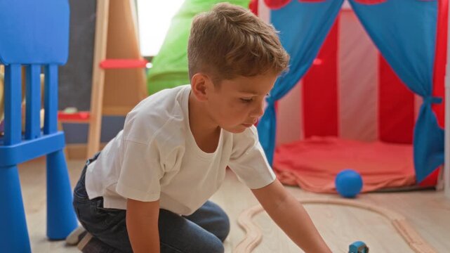 Boy pushing a wooden toy train along a curved track with hand on floor beside a red and blue play tent inside a building; curiosity focus.