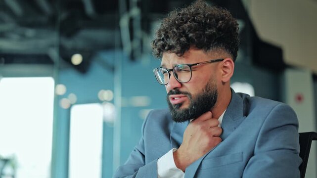 Middle Eastern businessman in a suit touching his neck in pain due to a sore throat. Image depicting illness, health problems, discomfort, and stress during a workday at the office.