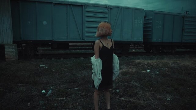 Young woman standing beside freight train on desaturated dusk railway