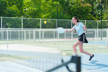 Obraz premium Dynamic action shot of young caucasian woman in blue athletic wear swinging pickleball paddle to hit the yellow ball in modern outdoor court on sunny day. Sports, healthy and active lifestyle concept