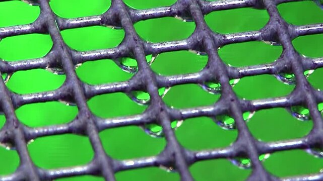 Magnified view of a hydro filter grid reveals countless water droplets clinging to its dark surface. A vibrant emerald green background glows softly behind the intricate mesh.