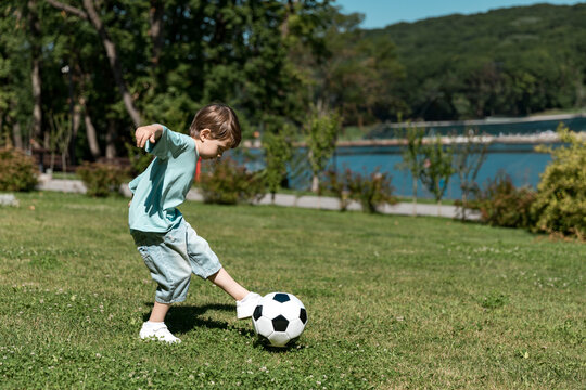 Side view little boy wearing blue shirt and shorts kicks a black and white soccer ball on green grass, with lake and trees background. Kids sport, childhood concept. Summer holiday camp. Copy space