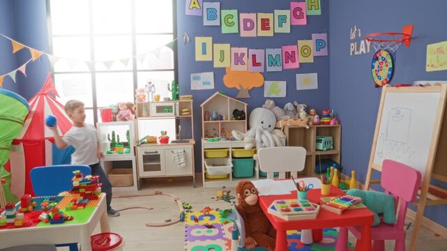 Boy holding blue ball walking through a playroom strewn with toys in building; childhood play joy memory.