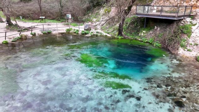 Aerial tracking shot pulling back over the deep circular pool of the Blue Eye spring, revealing the vibrant turquoise mineral water, green algae, and viewing platform in Albania