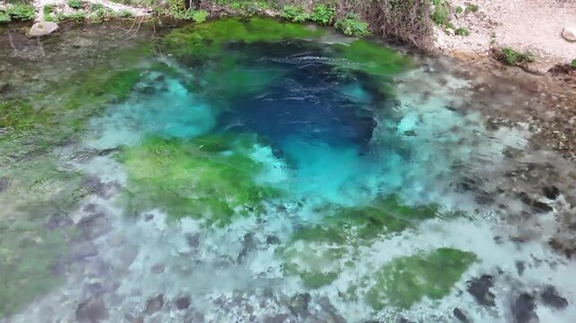 Aerial tracking shot rising vertically over the vibrant circular pool of the Blue Eye spring, revealing layers of deep blue water, bright turquoise rings, and bright green moss in Albania