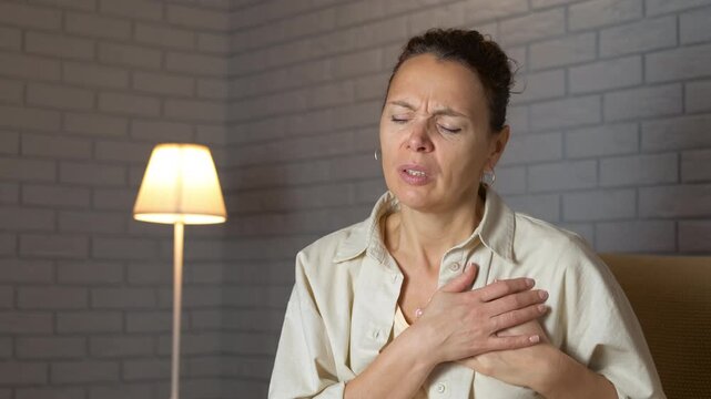 Woman experiencing sharp chest pain and heart attack symptoms. Middle-aged woman sitting at home suddenly feeling an intense chest pain, holding her heart and grimacing from discomfort