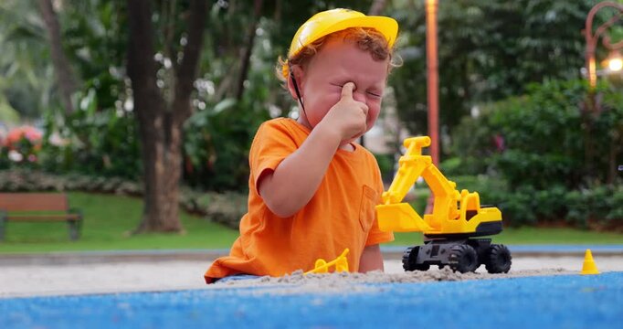 Toddler boy plays in sandbox when sand suddenly gets in his eyes, causing distress. He rubs his irritated eyes, ready to cry, and calls for his mother. She quickly arrives, wipes his eyes and calms