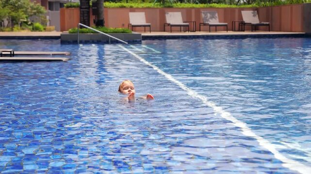 In shallow half of swimming pool on sunny, hot day, toddler boy lies in water with only his head and feet above surface. Relaxing and playing, he enjoys cool water, soaking in summer heat.