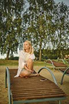 A woman sits comfortably on a lounge chair at the beach as the warm sunlight illuminates her surroundings. Trees sway gently in the breeze, creating a peaceful atmosphere during the evening