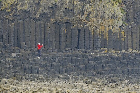 Person with red jacket standing in front of impressive basalt columns on a rocky cliff, Staffa, Mendelsons Hebrides Overture, Inner Hebrides, Scotland, United Kingdom