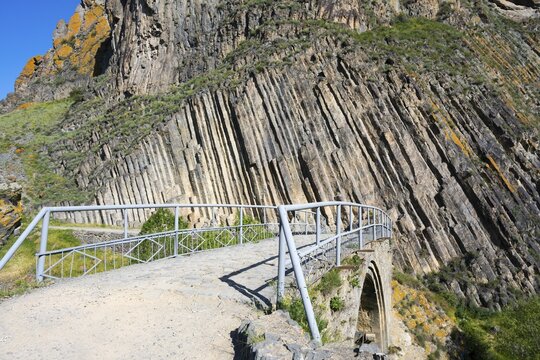 A narrow path with railing leads over a stone bridge, surrounded by unique vertical rock formations, Melik Tangi Bridge, behind it a basalt column formation, Vorotan River, Syunik Province, Armenia