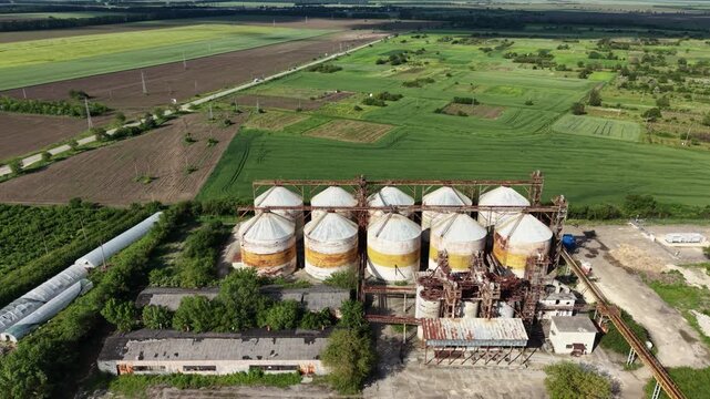 Aerial view of industrial grain silos with rusty metal roofs surrounded by lush green agricultural fields and trees in Balchik, Dobrich Province, Bulgaria.