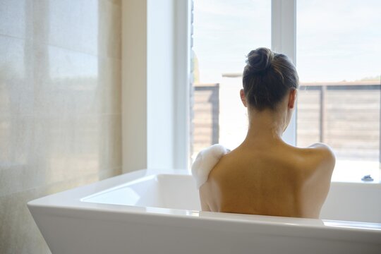 A person enjoys a calming bath in a contemporary bathroom filled with soft natural light. The atmosphere is peaceful as the individual leans back, surrounded by minimalist decor and privacy